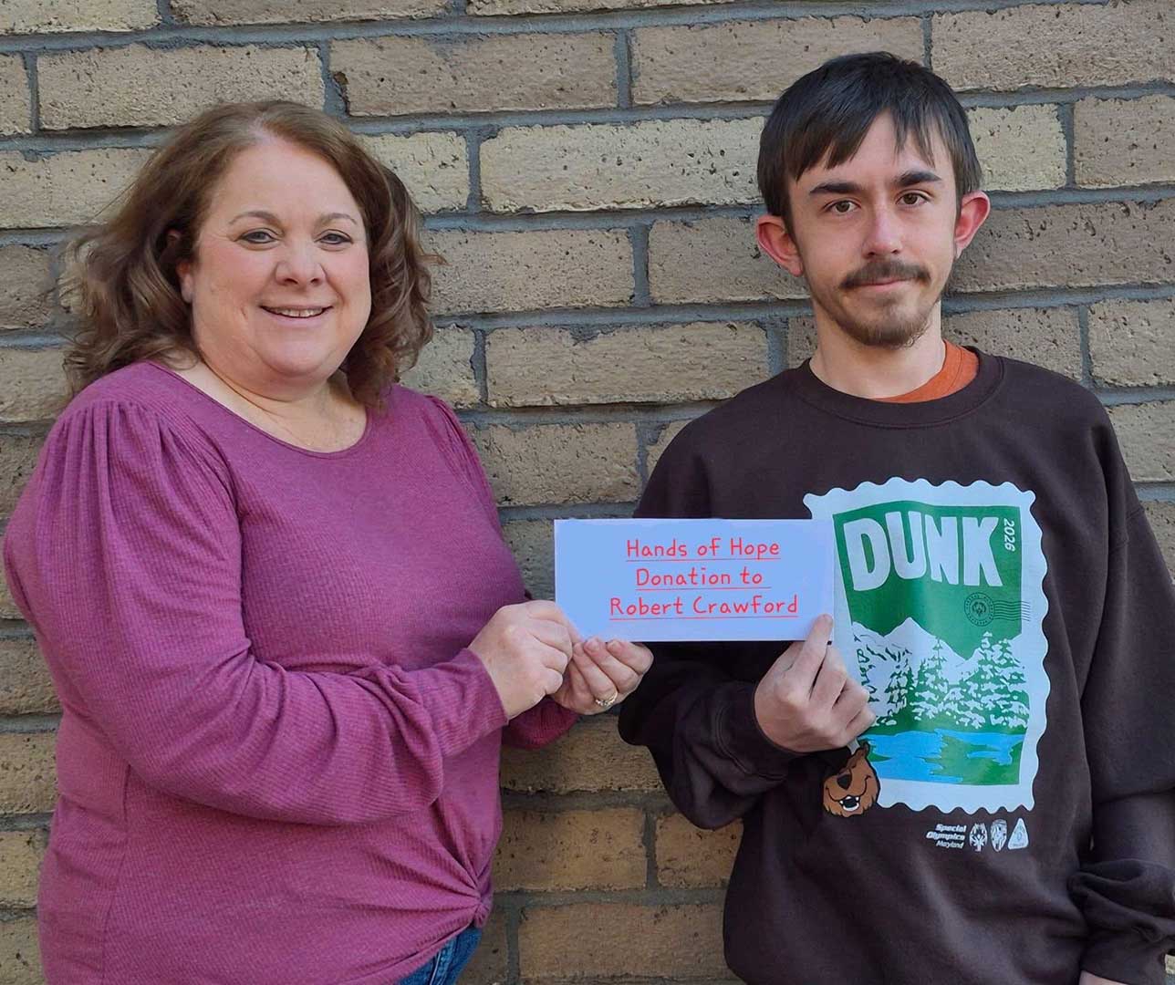 A smiling woman and a young man stand together in front of a brick wall, holding a card that reads "Hands of Hope Donation to Robert Crawford."