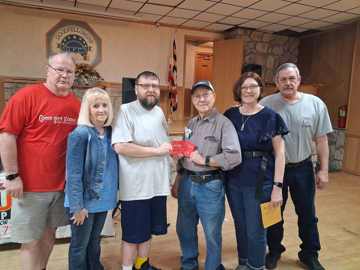 Six people pose for a group photo in a community hall, with two men in the center holding a red card that mentions a donation